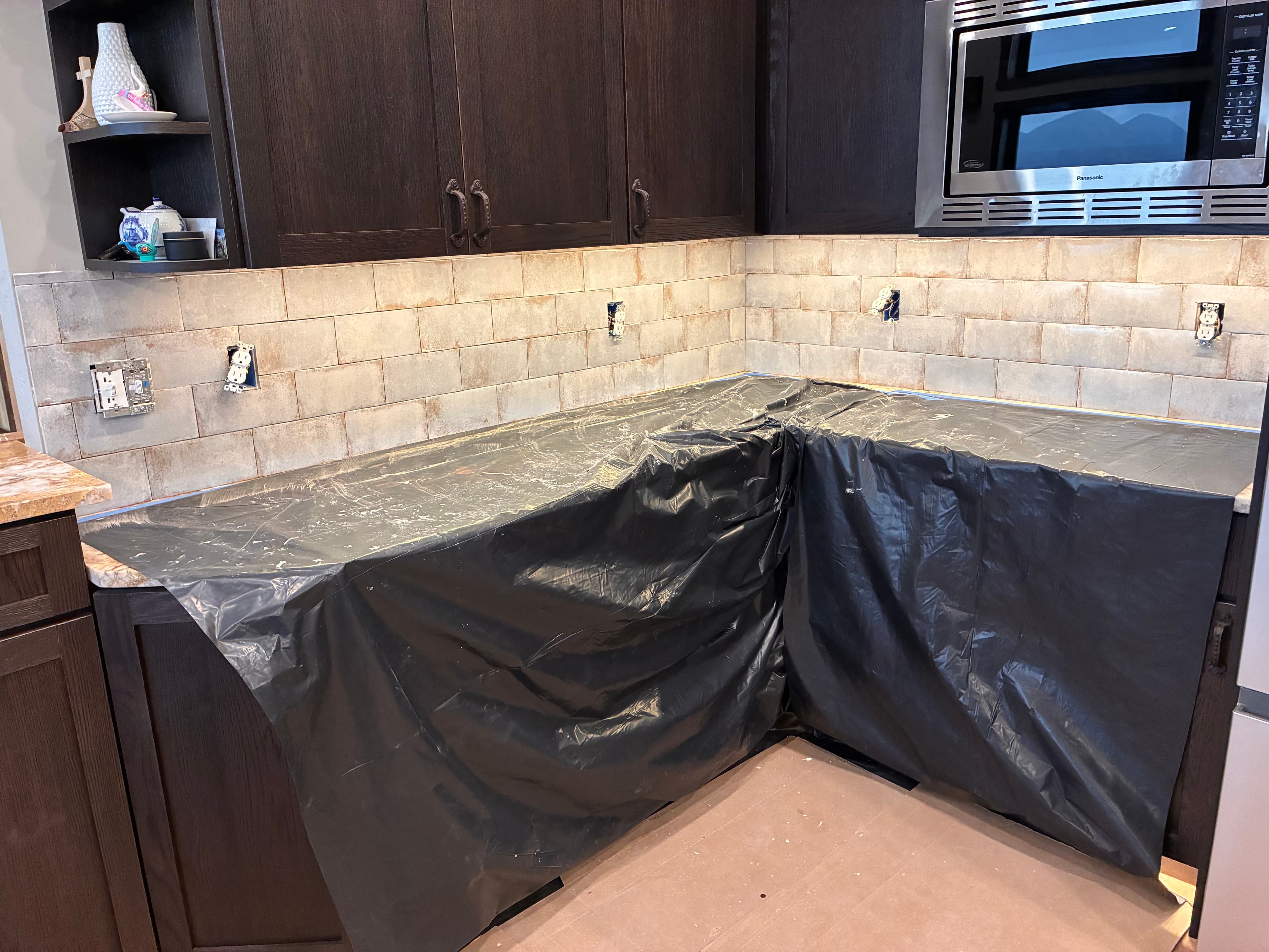 Kitchen renovation with dark cabinets, new tile backsplash, and countertops covered in black plastic.