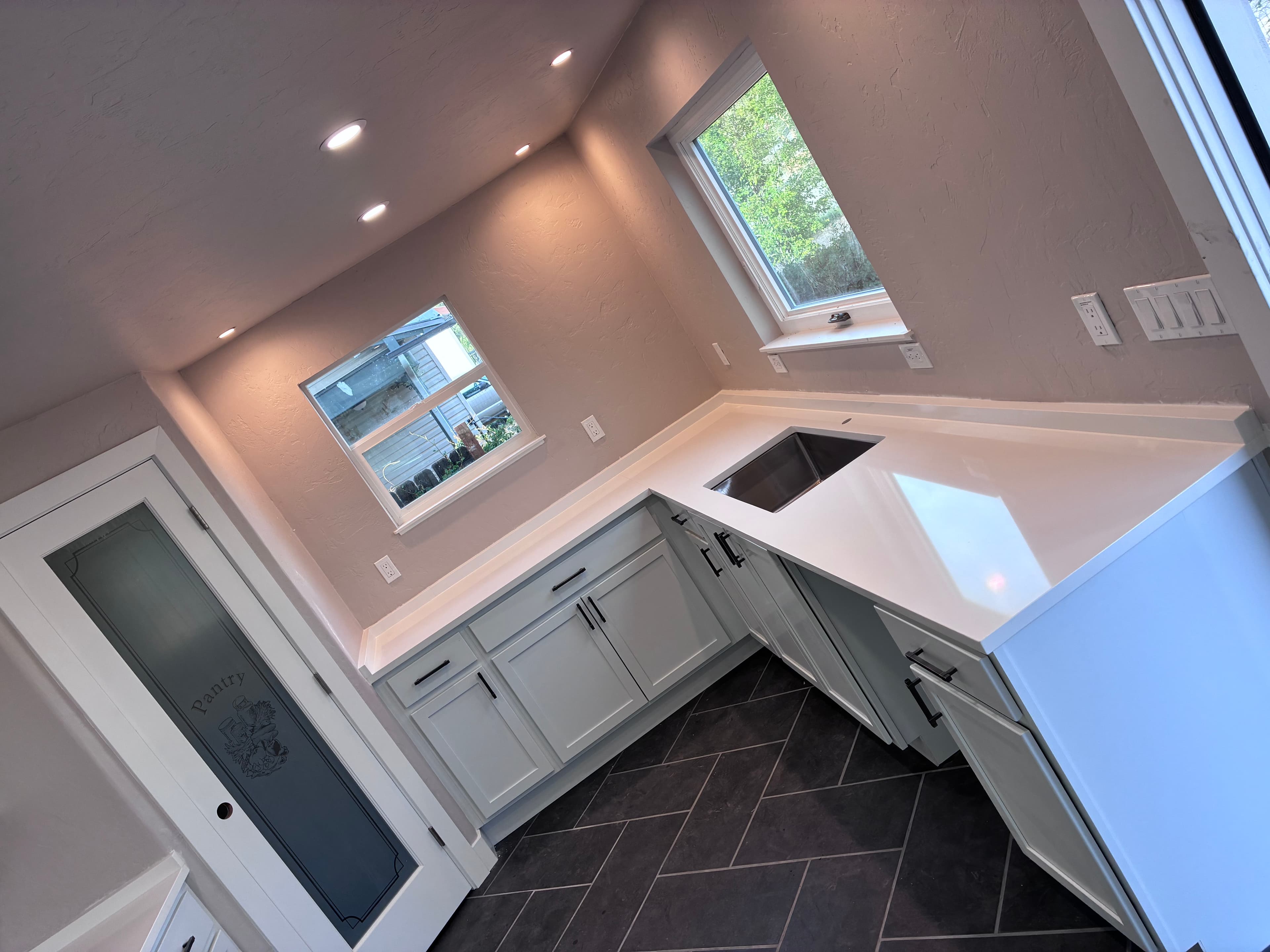 Modern kitchen featuring white cabinets, white countertops, dark herringbone tile floors, and a pantry door.