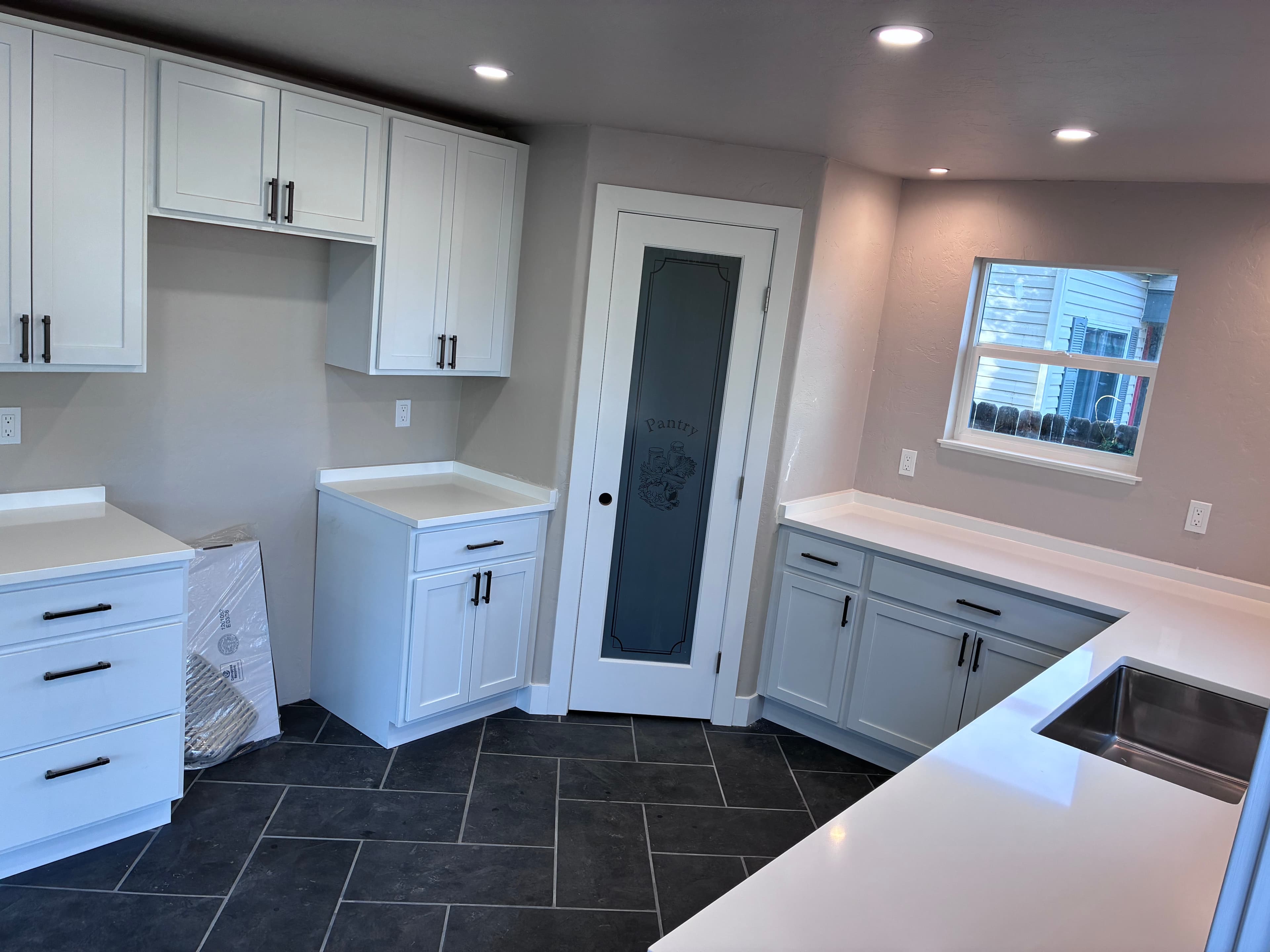 Modern kitchen with white cabinets, black hardware, dark tile floors, and frosted glass pantry door.