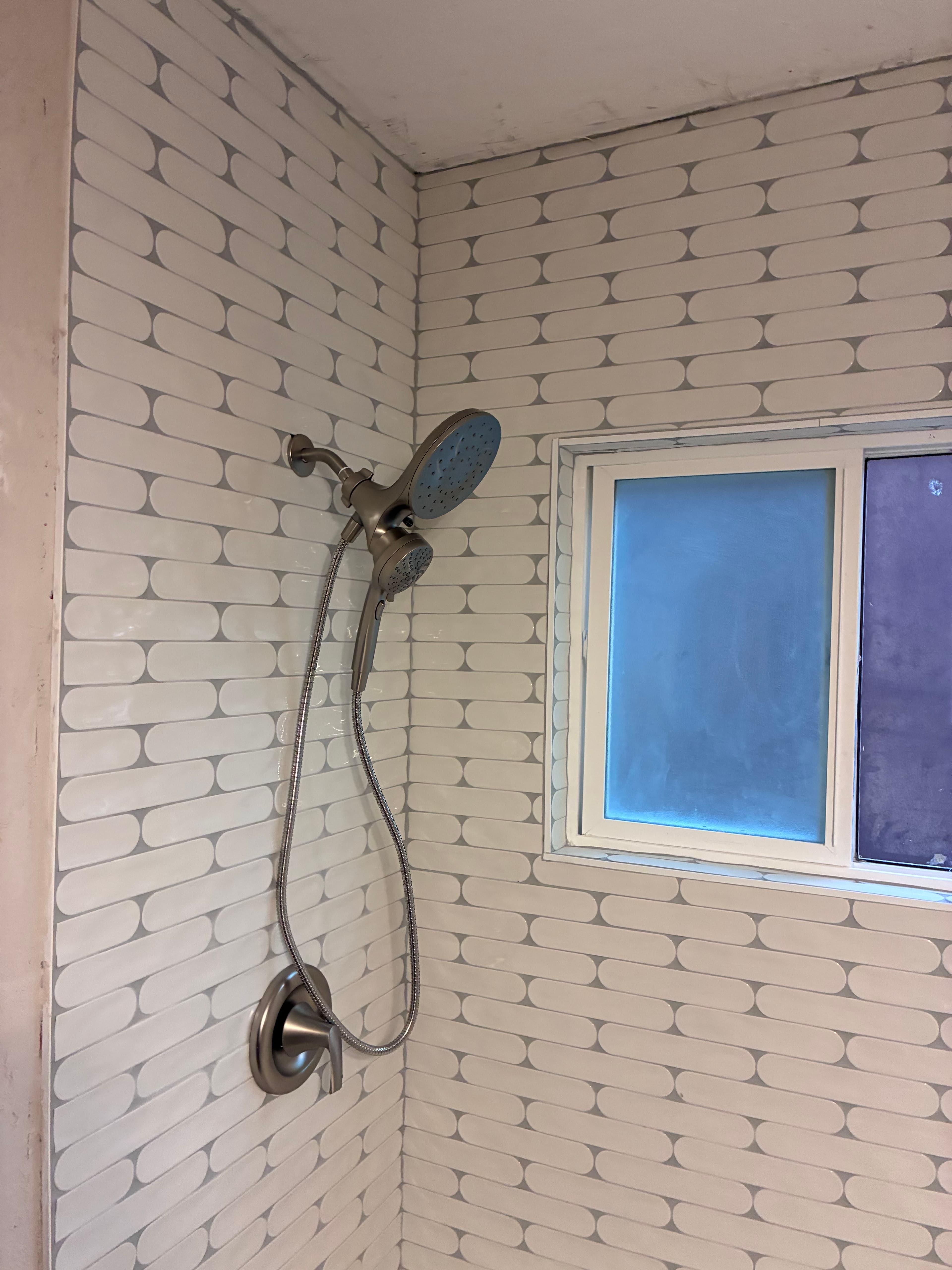 Brushed nickel dual shower head on white pill-shaped tile wall beside a frosted window.